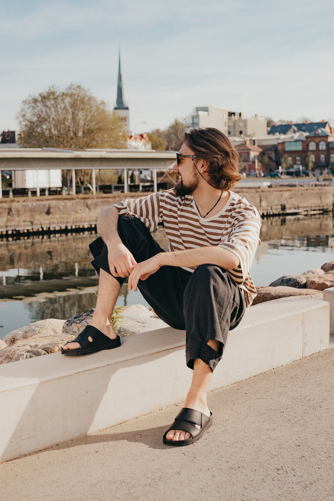 1. Men's black Miku sandals by Omaking worn by model sitting outdoors by the water