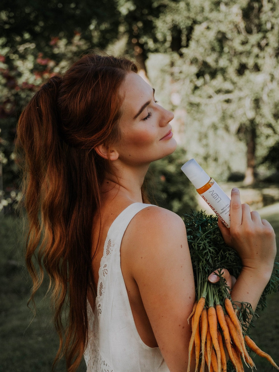 1. Woman lying on striped towel with LUUV Carrot Dry Oil and other skincare products, surrounded by flowers and a wicker basket