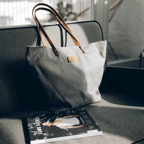 5. Grey denim tote bag by Muni with tan leather handles, placed on grey sofa with magazine in lifestyle setting