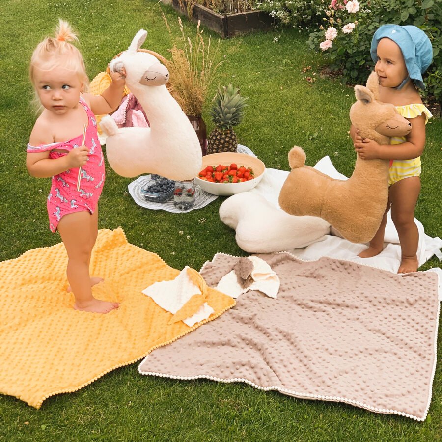 2. Two children playing with cream and brown alpaca soft toy-pillows on colorful blankets in a garden