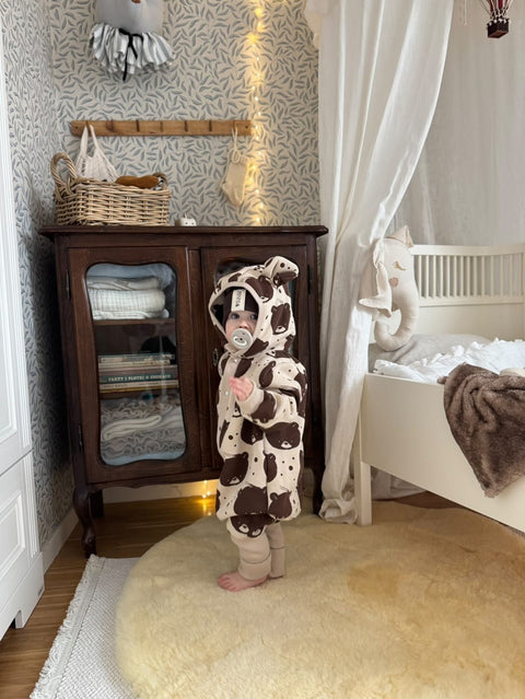 1. Baby in beige bear-patterned jumpsuit with hood standing in a cozy nursery