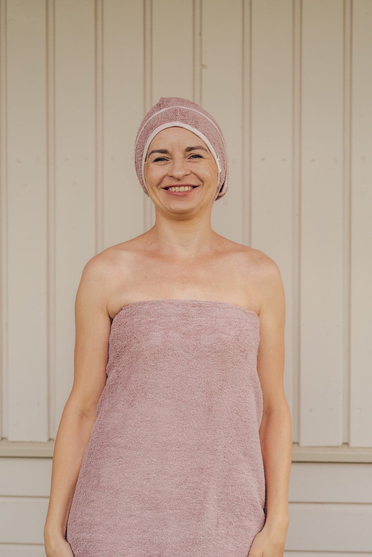 2. Woman smiling in RÄTT cotton terry hair wrap and towel set in pink, front view, lifestyle setting