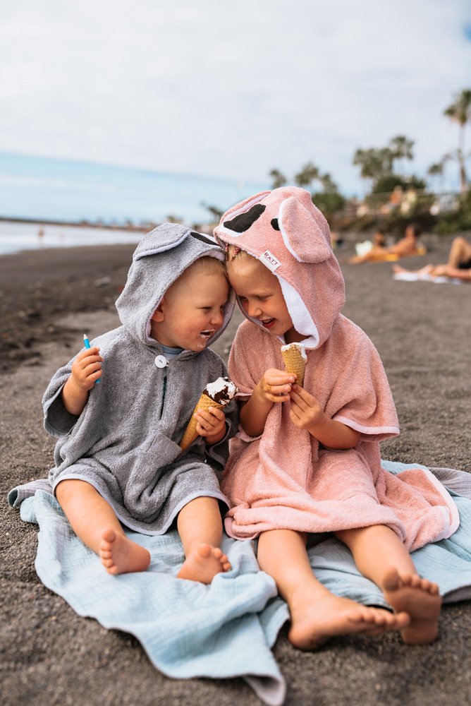 1. Toddlers wearing RÄTT short-sleeve ponchos in grey and pink, enjoying ice cream on the beach