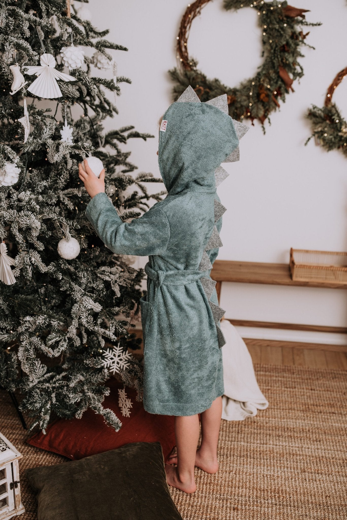 1. Child wearing RÄTT's green dinosaur-themed bamboo terry bathrobe with hood, standing by a decorated Christmas tree
