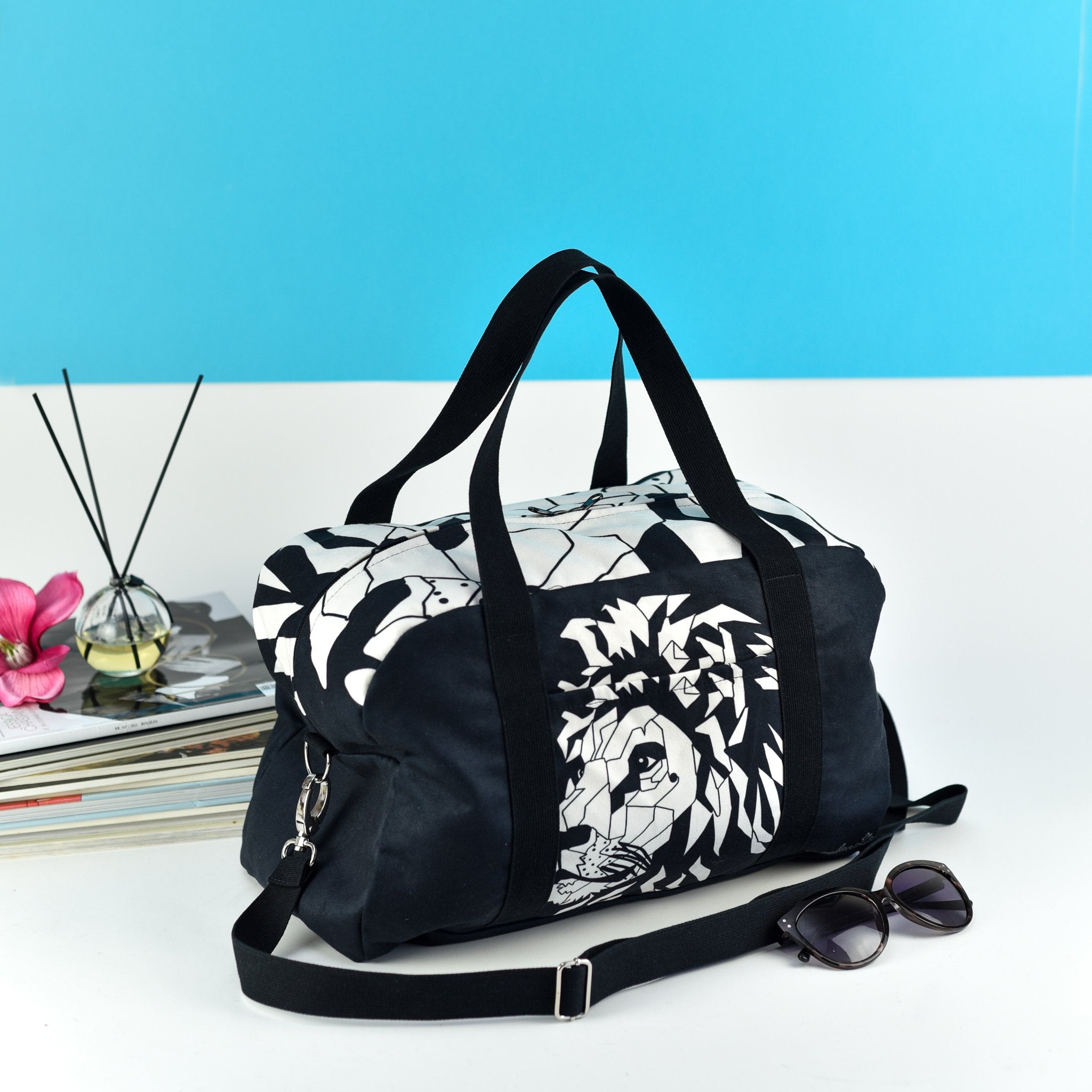 1. Garbanota Travel Bag White Lion in black with geometric lion design, shown in lifestyle setting with sunglasses and magazines