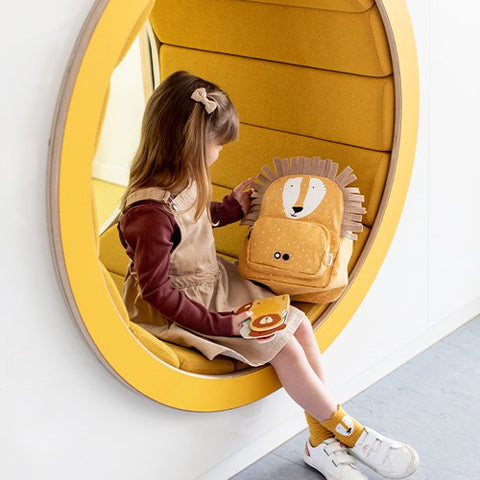 1. Young girl sitting in yellow nook holding Trixie Baby Mr Lion backpack, wearing beige dress and socks, creating a playful setting