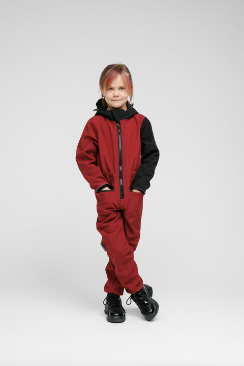 1. Child wearing Nuckö sunset red softshell onesie with black hood and sleeves, standing with arms crossed in studio setting