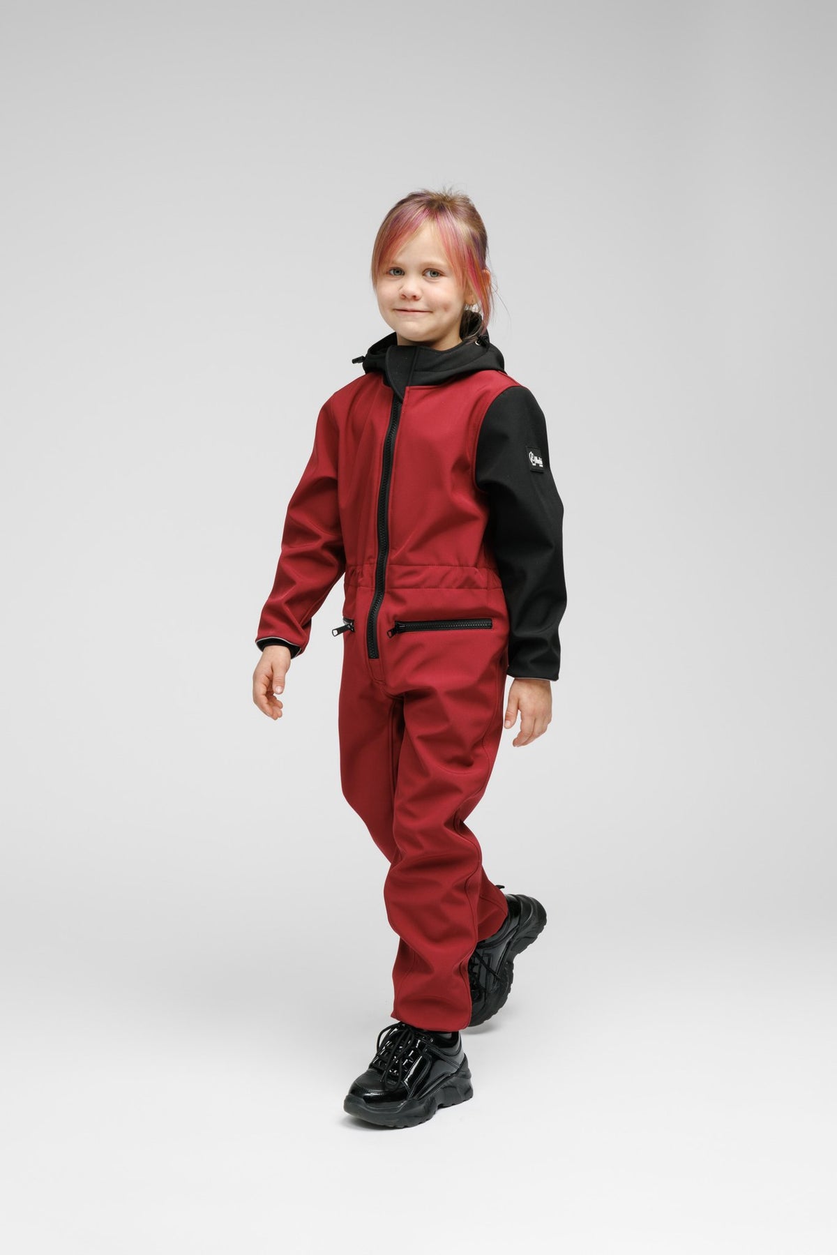 2. Child walking in Nuckö sunset red softshell onesie with black hood and sleeves, showing front zip and pocket details