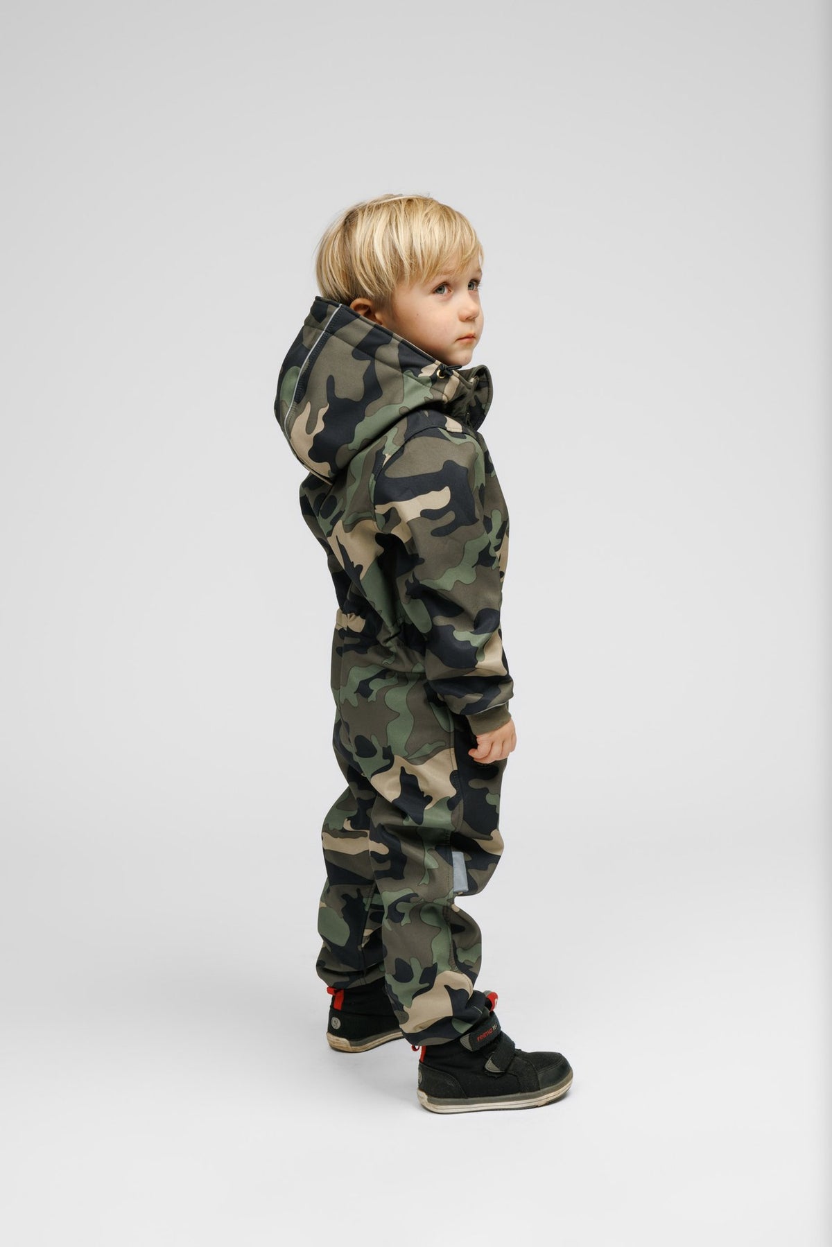 3. Side profile of child in Nuckö camouflage softshell onesie, showing zippered pockets and ribbed cuffs, studio background