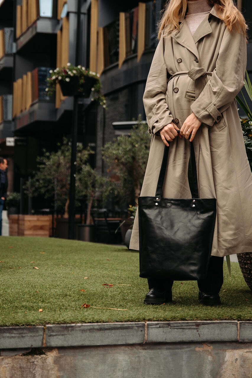 2. Woman standing on grass holding black leather tote bag by Stella Soomlais, wearing a beige trench coat in an urban setting