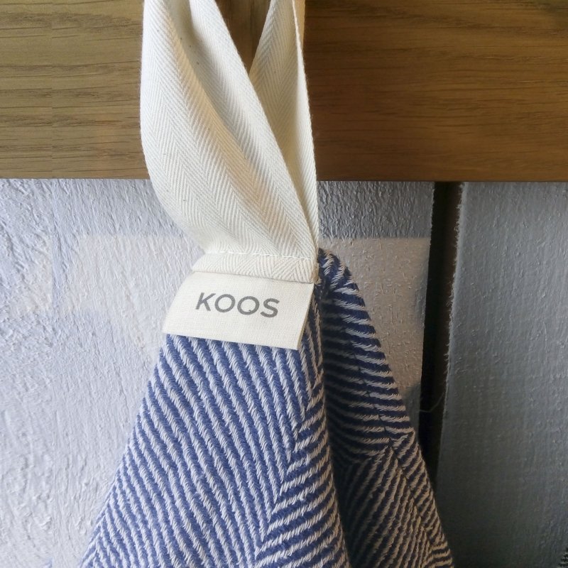 6. Blue linen towel with fishbone pattern hanging from wooden hook, showing KOOS label