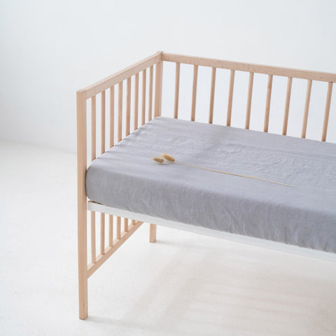 1. Light grey linen fitted sheet on a wooden crib in a minimalist room, showcasing the soft texture and elastic hem