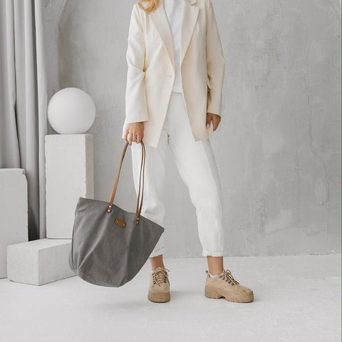 2. Grey denim tote bag by Muni with tan leather handles, worn by woman in white outfit, shown in lifestyle setting