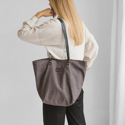 1. Woman carrying Muni brown denim tote bag with black leather handles, shown from the back in a lifestyle setting