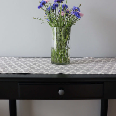 1. Table runner with Hanikatsi fly pattern on dark table, featuring a vase of blue and pink flowers, showcasing linen-cotton blend fabric