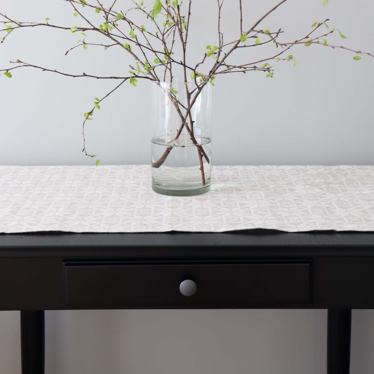 1. Beige table runner with kaheksakanna pattern on dark table, glass vase with branches, minimalist setting