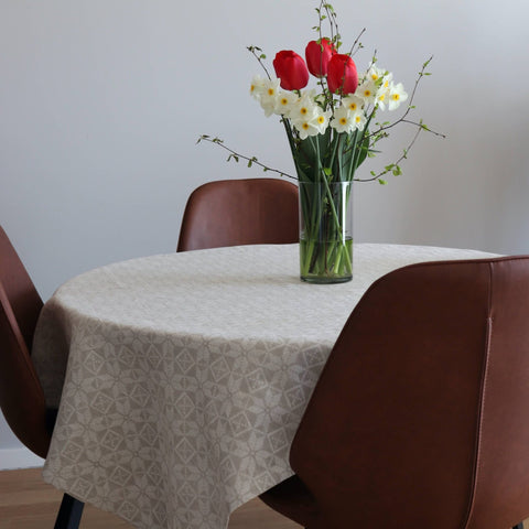 1. Beige tablecloth with Hiiumaa kaheksakanna mitten pattern on round table, decorated with vase of red and white flowers, surrounded by brown chairs in a modern dining room setting