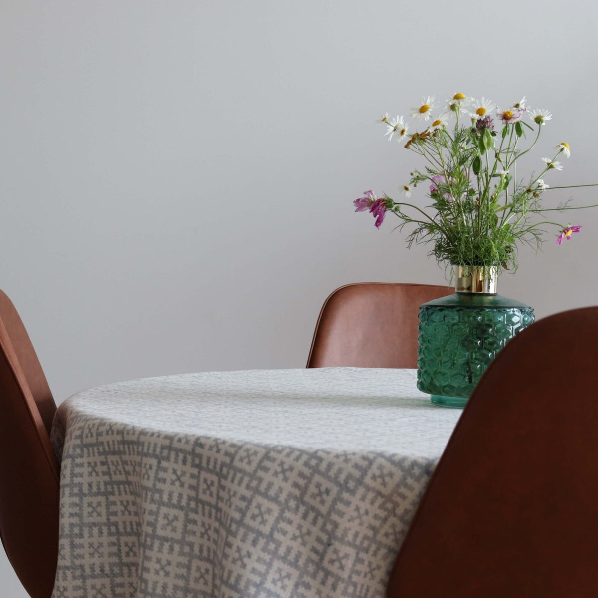 1. Round table with LOKO Setumaa tablecloth featuring traditional mitten pattern, adorned with a green vase of wildflowers, surrounded by brown chairs in a minimalist dining room