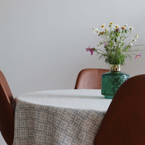 1. Round table with LOKO Setumaa tablecloth featuring traditional mitten pattern, adorned with a green vase of wildflowers, surrounded by brown chairs in a minimalist dining room