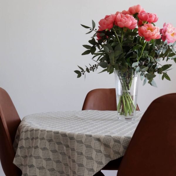 1. Table with LOKO Virumaa tablecloth featuring geometric pattern, adorned with pink flowers in a vase, surrounded by brown chairs in a dining setting