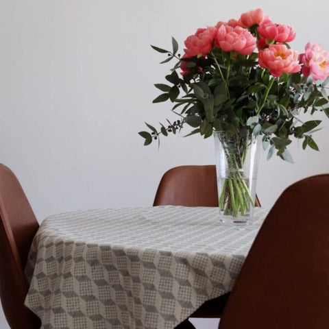1. Table with LOKO Virumaa tablecloth featuring geometric pattern, adorned with pink flowers in a vase, surrounded by brown chairs in a dining setting