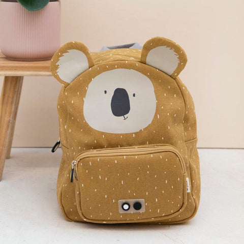 1. Trixie Baby Mr Koala backpack in mustard yellow with koala face, placed next to a pink plant pot on a wooden stool