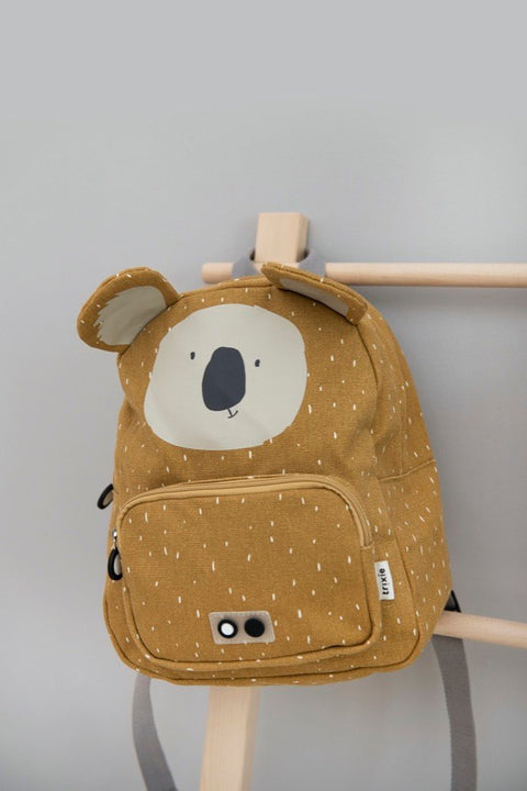 1. Trixie Baby Mr Koala backpack in mustard yellow with koala face, hanging on wooden rack in a room setting