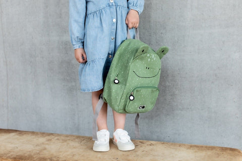 1. Child holding green Mr Frog backpack by Trixie Baby, wearing a blue dress and white sneakers, standing against a concrete wall
