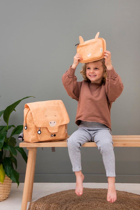 1. Child sitting on bench holding Trixie Baby Mrs Giraffe school bag in orange, smiling in indoor setting