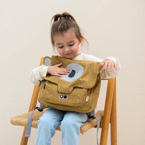 1. Young girl sitting on chair holding Trixie Baby Mr Koala school bag in mustard yellow, smiling and interacting with the bag