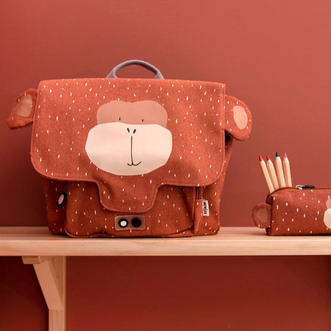 1. Trixie Baby Mr Monkey school bag on wooden shelf with matching pencil case, set against a warm brown wall