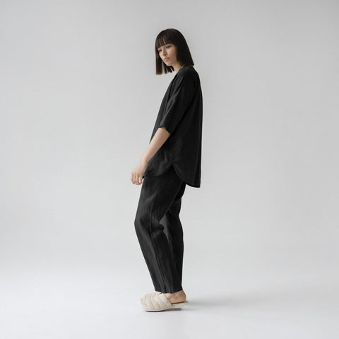 1. Woman wearing black linen loungewear set with relaxed fit, V-neck top, and 3/4-length sleeves, standing in side profile on a plain background