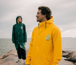 1. Unisex Moomin Adventure Rain Jacket in dark green and yellow worn by male and female models on rocky shore