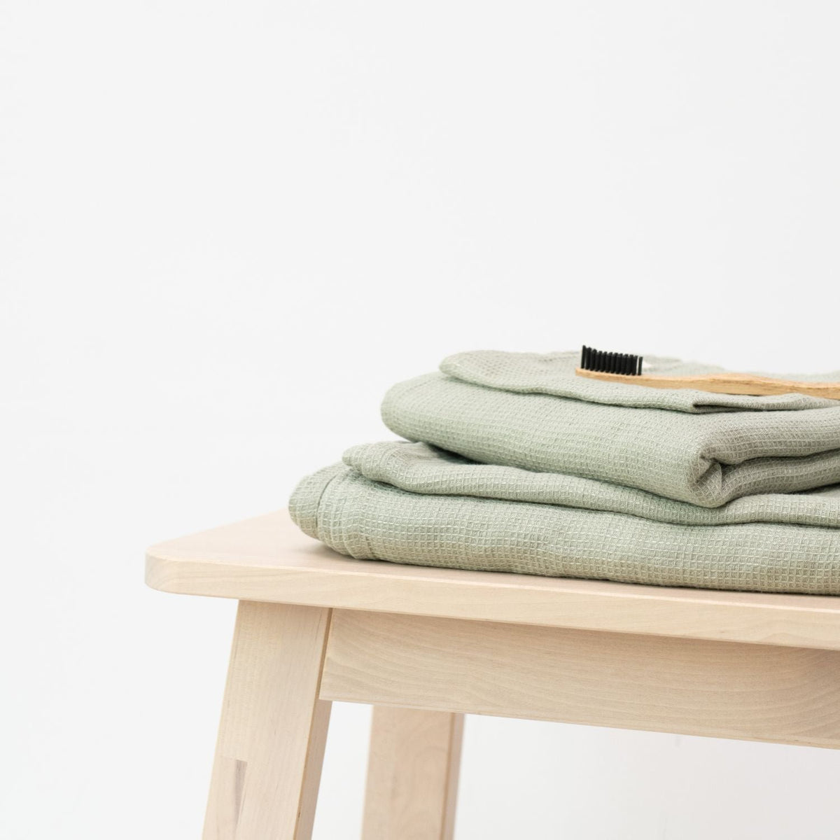 2. Sage green waffle linen towels neatly folded on a wooden stool with a grooming brush