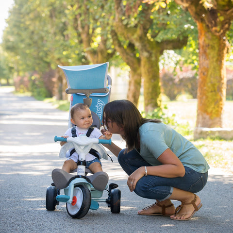 1. Woman crouching next to a child on a blue Smoby Baby Balade Plus tricycle in a sunny park