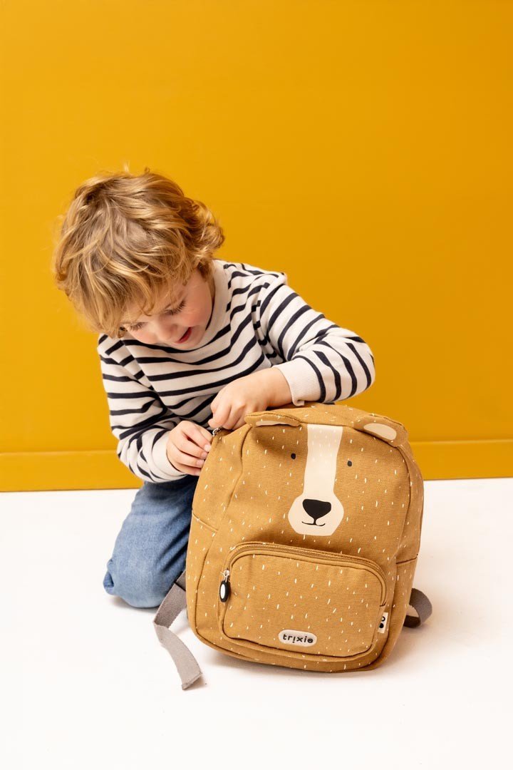 1. Child playing with Trixie Baby Mr. Bear backpack, kneeling against a yellow background