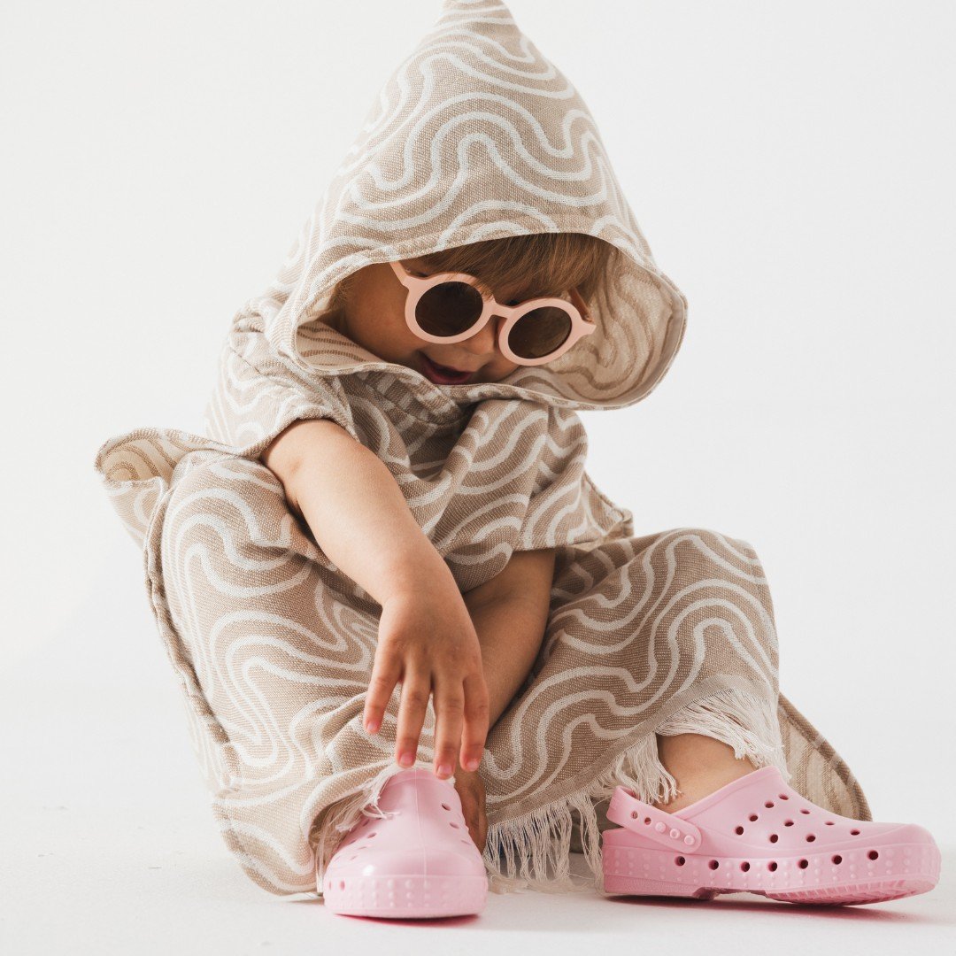 3. Child in sand-colored bamboo bath poncho with hood and pink sunglasses reaching for pink shoes