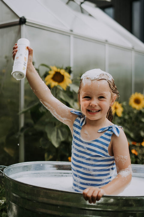 1. Smiling child in striped swimsuit holding LUUV Caring Kid's Shampoo Vanilla bottle in outdoor bath with sunflowers
