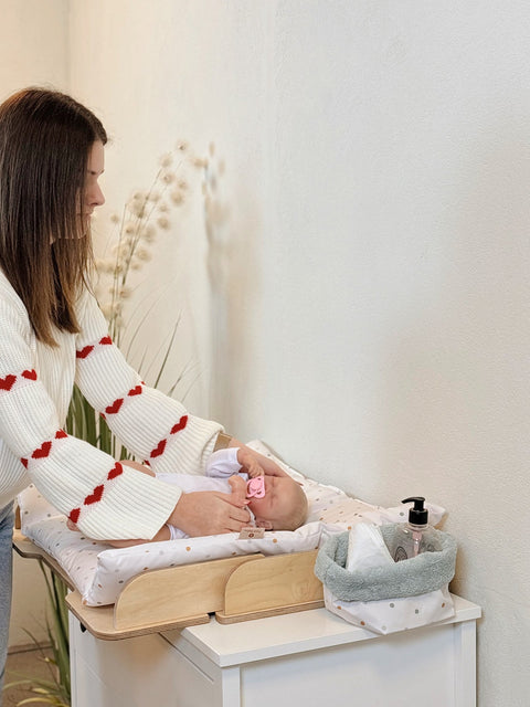 3. Woman in white sweater with red hearts changing a baby on a polka dot mat with a grey storage basket nearby