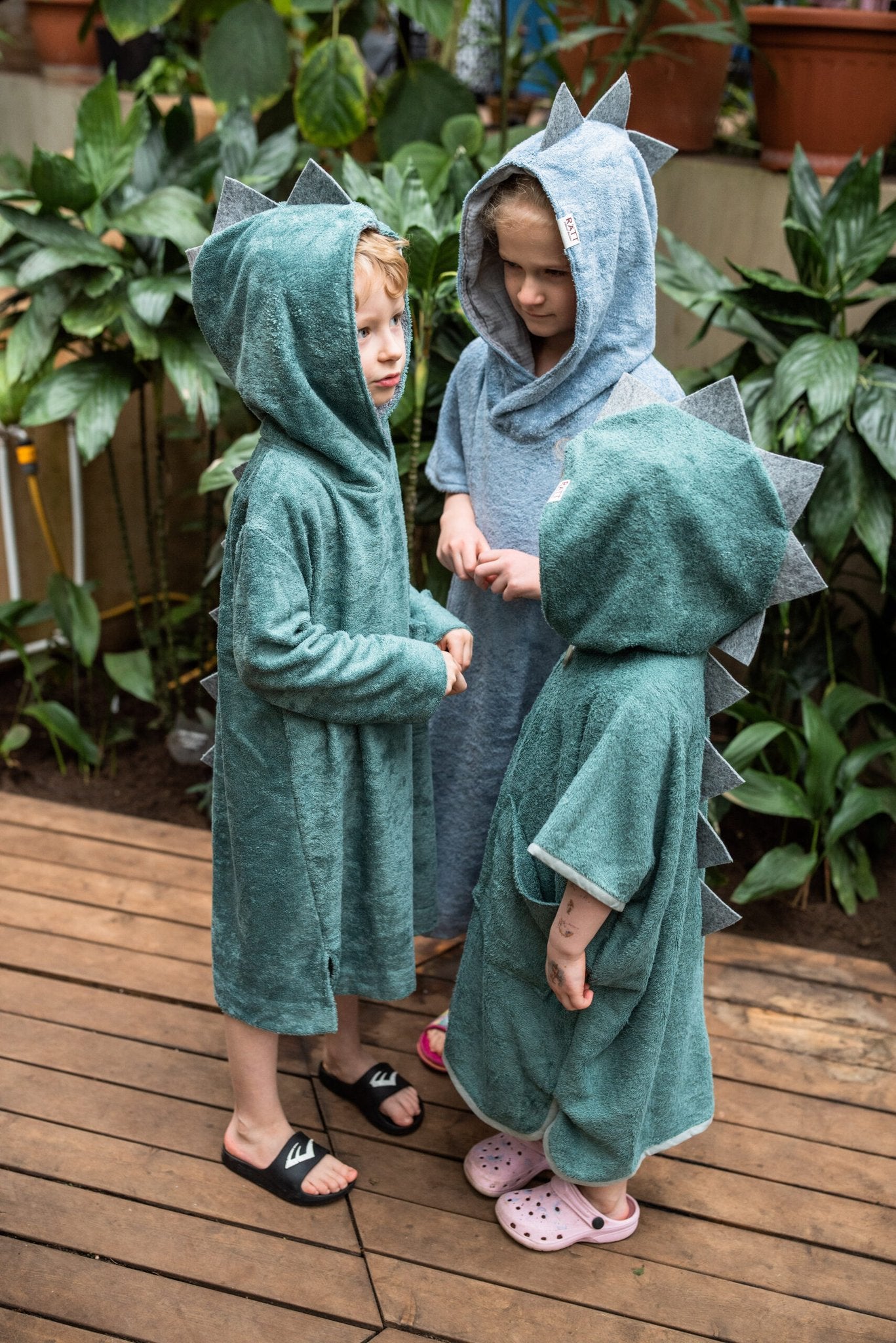 1. Toddlers wearing RÄTT long-sleeve dinosaur ponchos in green and blue, shown in a garden setting