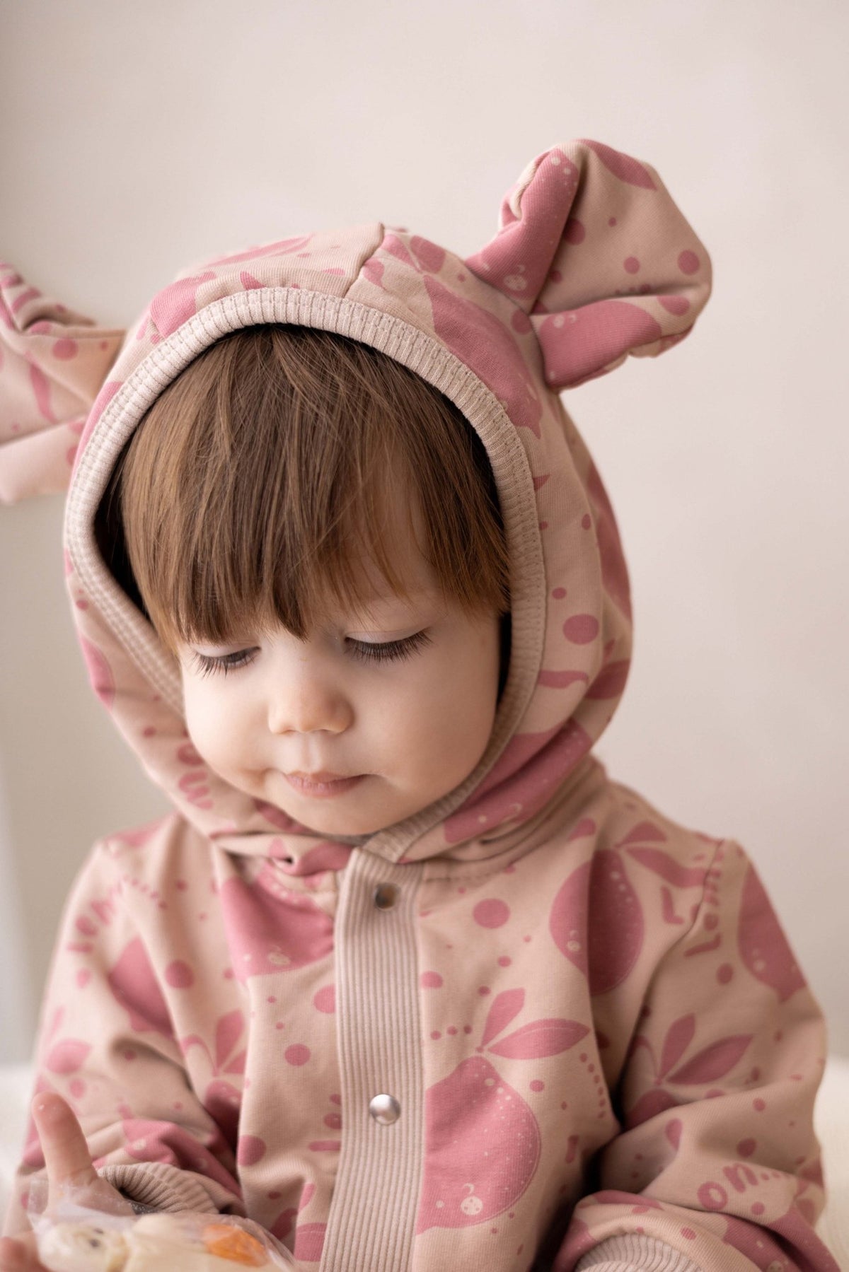 1. Baby looking down wearing Zezuzulla beige jumpsuit with pink lemon print and hood with ears indoors