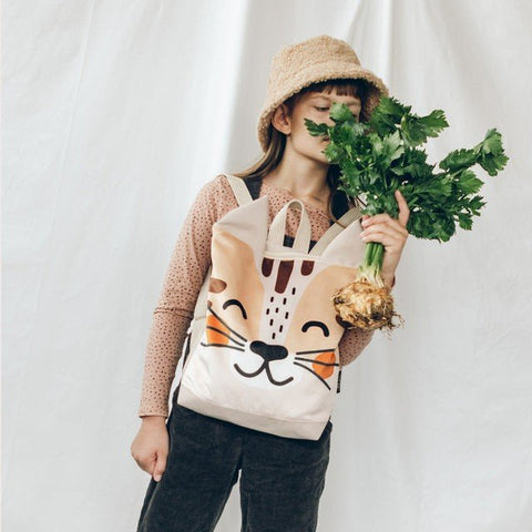 1. Girl wearing MUNI cat backpack holding celery, dressed in pink top and hat, against white backdrop