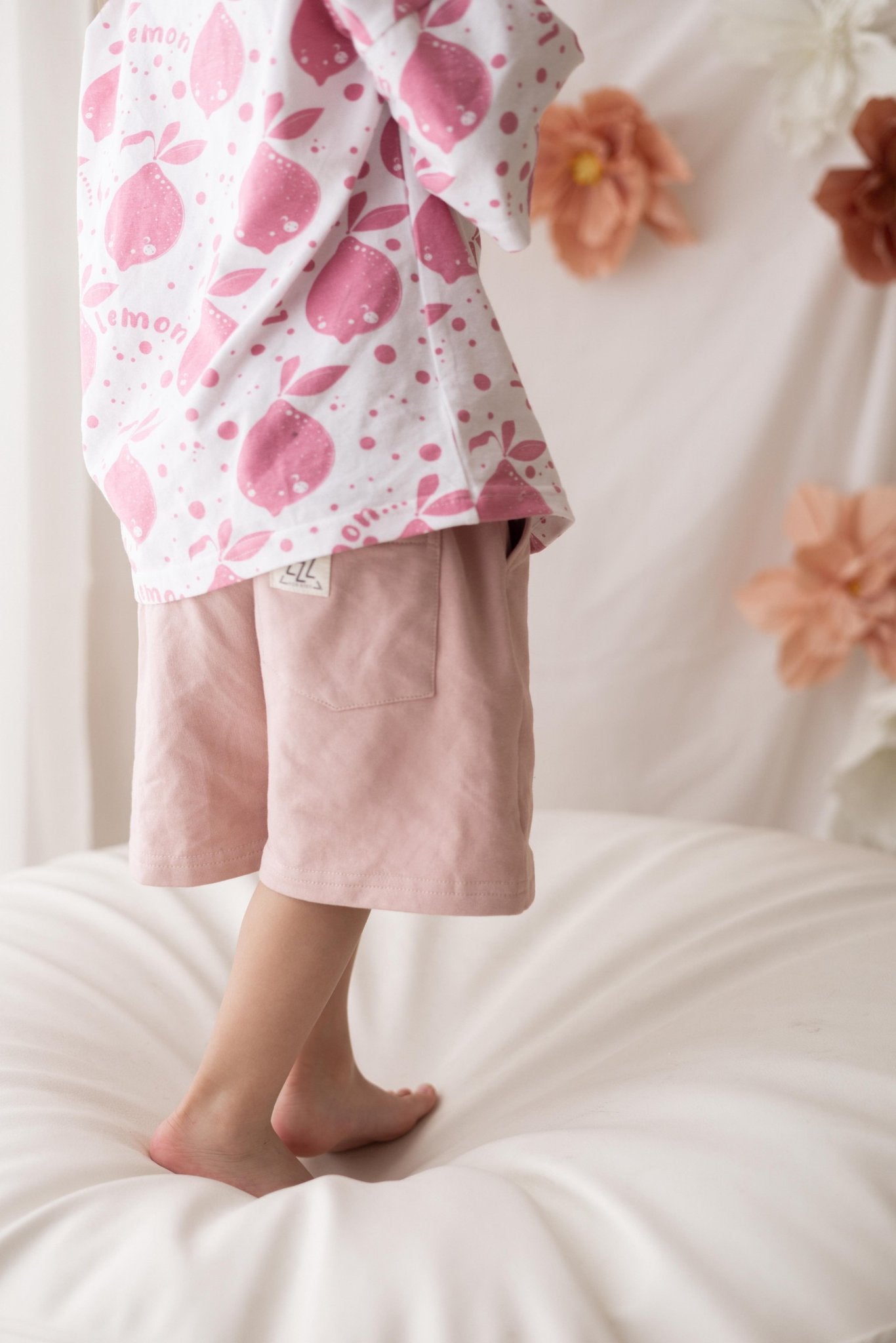 1. Child wearing pastel pink shorts with elastic waistband and white printed shirt standing on a soft surface with floral background