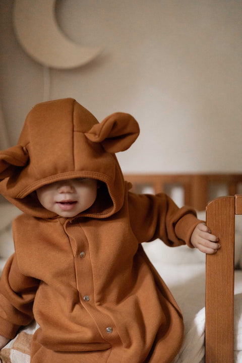 1. Baby in cinnamon eared jumpsuit with hood, standing in crib with moon decor in background