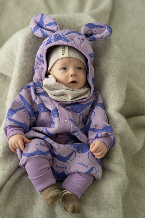 1. Baby wearing lilac Fenio jumpsuit with hood and ears, lying on a soft blanket