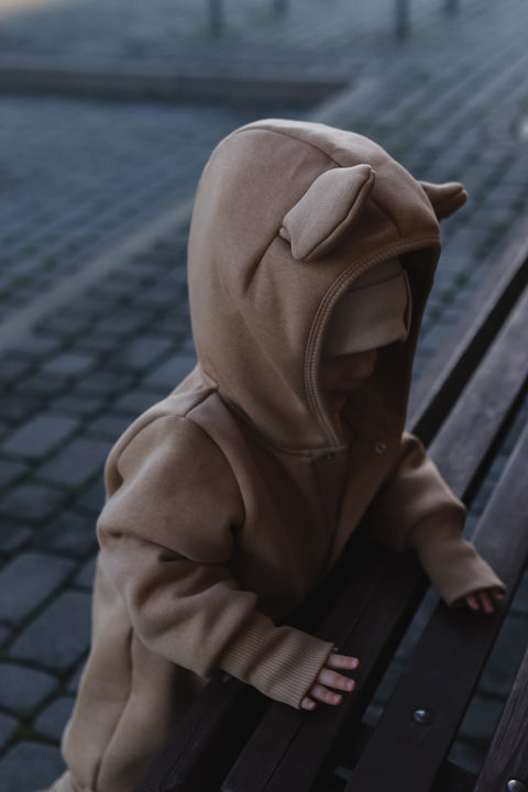 3. Close-up of baby in cacao eared jumpsuit with hood and ears, leaning on a bench