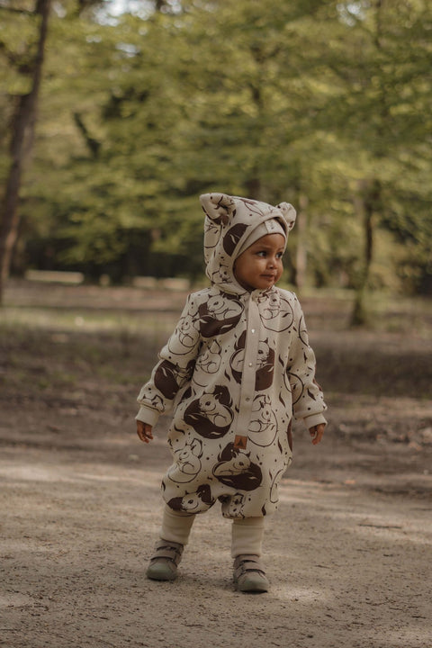 1. Child wearing beige squirrel print jumpsuit with hood and ears, standing on a dirt path in a forest