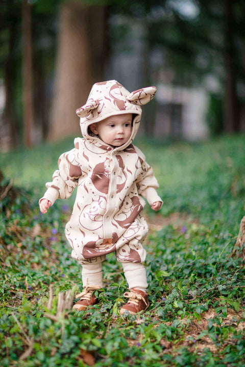 1. Baby wearing beige squirrel print jumpsuit with hood and ears, standing on grass in a park