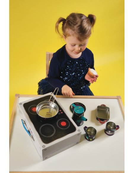 3. Young girl playing with space-themed tea set on a table, including a coffee pot, cups, and cooking utensils, against a yellow background