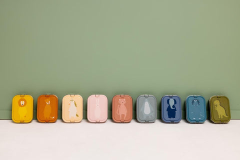 1. Lineup of Trixie Baby lunch boxes with different animal designs against green wall